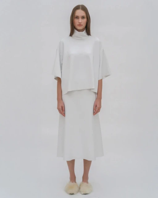 Studio shot of a model in a white turtleneck top and midi skirt, posing against a white background. She is wearing furry slippers.