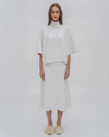 Studio shot of a model in a white turtleneck top and midi skirt, posing against a white background. She is wearing furry slippers.