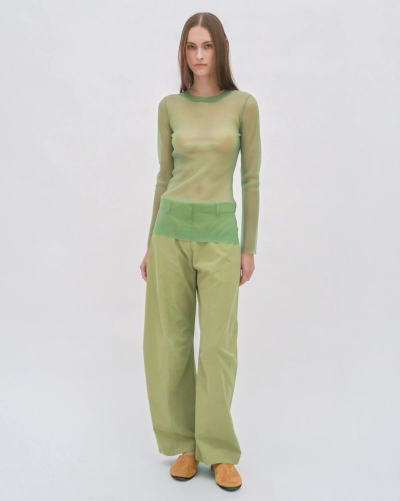 A woman in her twenties wearing a light green sheer top and baggy trousers standing in a white photo studio.