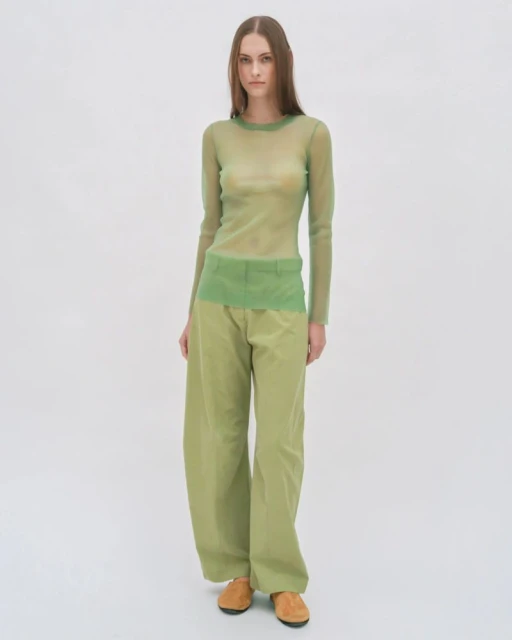 A woman in her twenties wearing a light green sheer top and baggy trousers standing in a white photo studio.