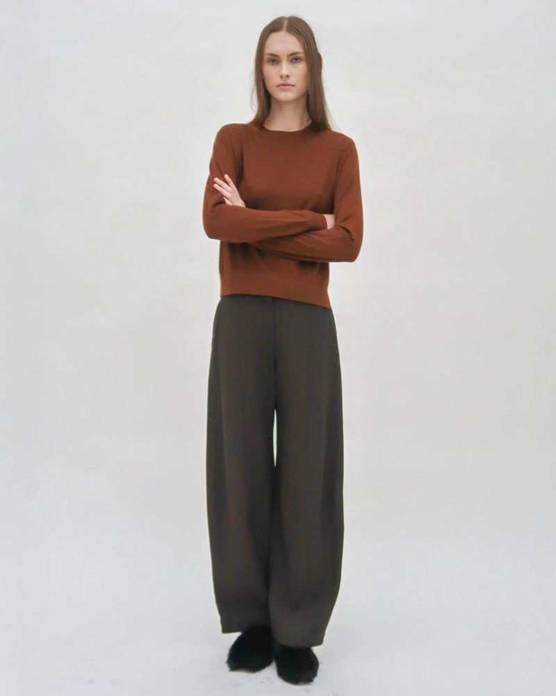 A female model standing with arms crossed wearing a brown sweater and dark wide-leg trousers against a neutral backdrop.