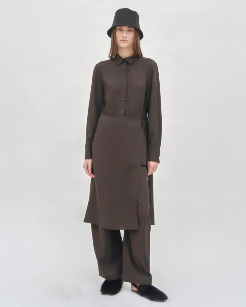 Studio shot of a female fashion model in a long-sleeved brown shirt dress, matching wide-leg pants, bucket hat, and fluffy slippers on a white backdrop.