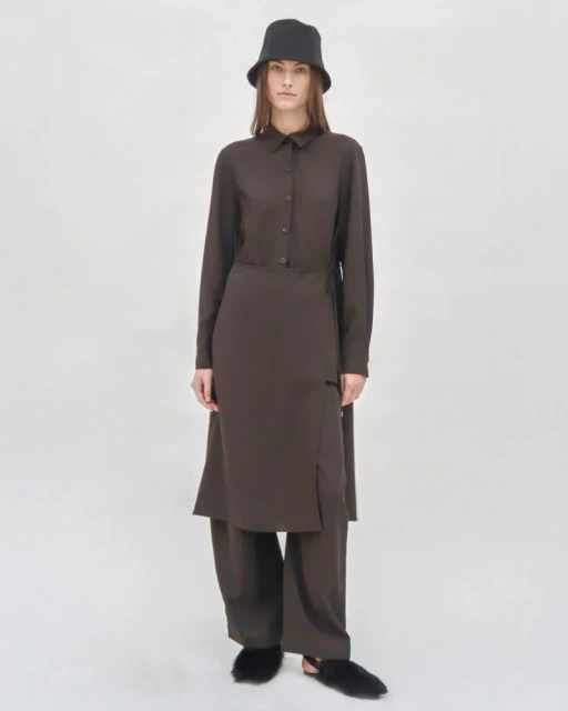 Studio shot of a female fashion model in a long-sleeved brown shirt dress, matching wide-leg pants, bucket hat, and fluffy slippers on a white backdrop.