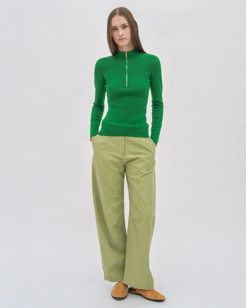 Model wearing a green zip-up turtleneck and light green wide-leg trousers against a white seamless backdrop