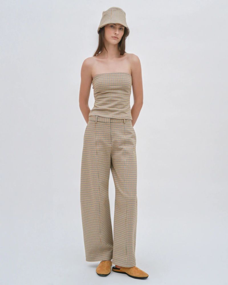 A woman poses in a studio wearing a tan, brown, and black plaid two-piece outfit, consisting of a strapless top, wide-legged pants, and a bucket hat.