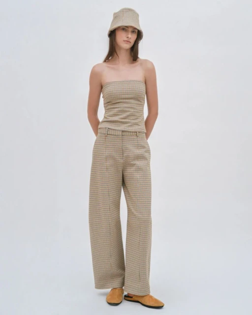 A woman poses in a studio wearing a tan, brown, and black plaid two-piece outfit, consisting of a strapless top, wide-legged pants, and a bucket hat.