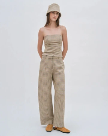 A woman poses in a studio wearing a tan, brown, and black plaid two-piece outfit, consisting of a strapless top, wide-legged pants, and a bucket hat.