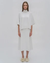 Model standing against a white backdrop wearing a white turtleneck top, midi skirt, and fluffy slippers for Rosetta Getty S/S 25.