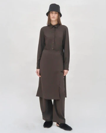Model in a brown shirt dress and trousers wearing a black bucket hat against a plain white studio backdrop
