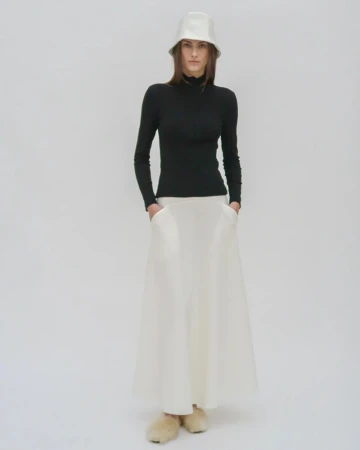 Model wearing a white bucket hat, black turtleneck, and white skirt against a plain light grey studio backdrop.