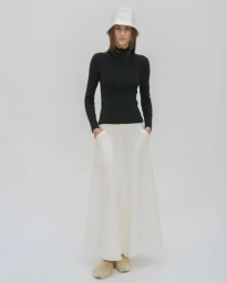 Model wearing a white bucket hat, black turtleneck, and white skirt against a plain light grey studio backdrop.