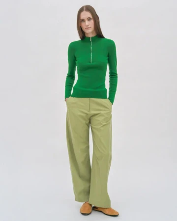 Model wearing a green zip-up turtleneck and light green wide-leg trousers against a white seamless backdrop