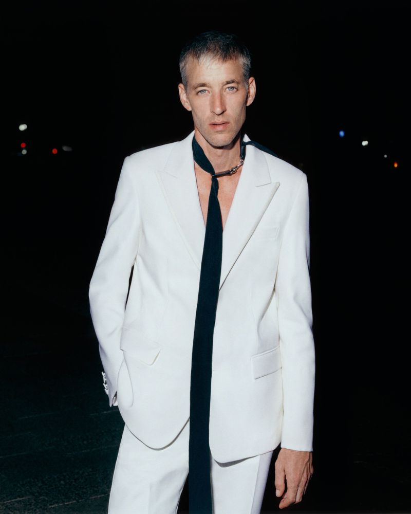 A man in a white suit and black tie stands outdoors at night, posing for a fashion portrait. The background is blurred and dark.