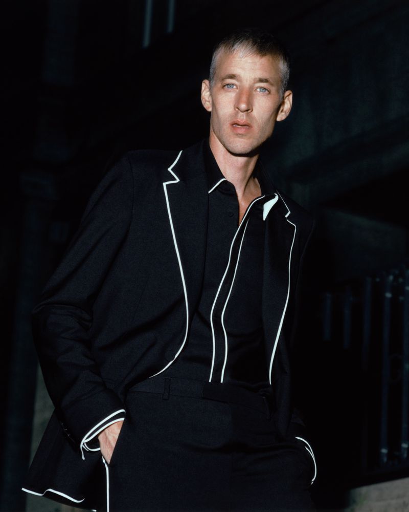 Man wearing a black suit with white piping details against a dark architectural exterior setting at night.