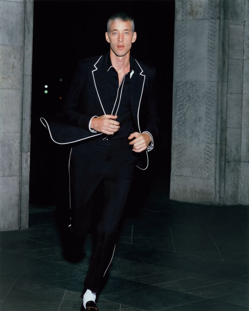 Adult male fashion model in a black suit with white trim running outdoors at night near an architectural stone structure.