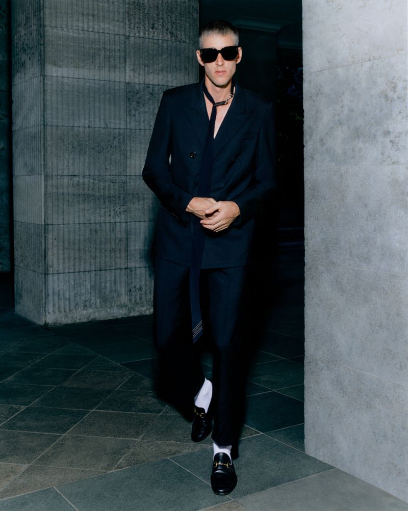 Man in suit and sunglasses walking on patterned tile. He wears a dark double-breasted suit, socks, and loafers in an indoor setting.