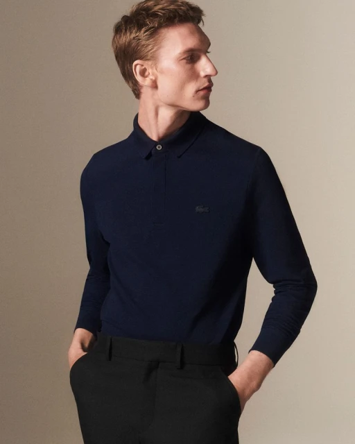 A fair-skinned, auburn-haired adult male poses indoors, wearing a long-sleeve navy polo shirt and black slacks against a neutral background.