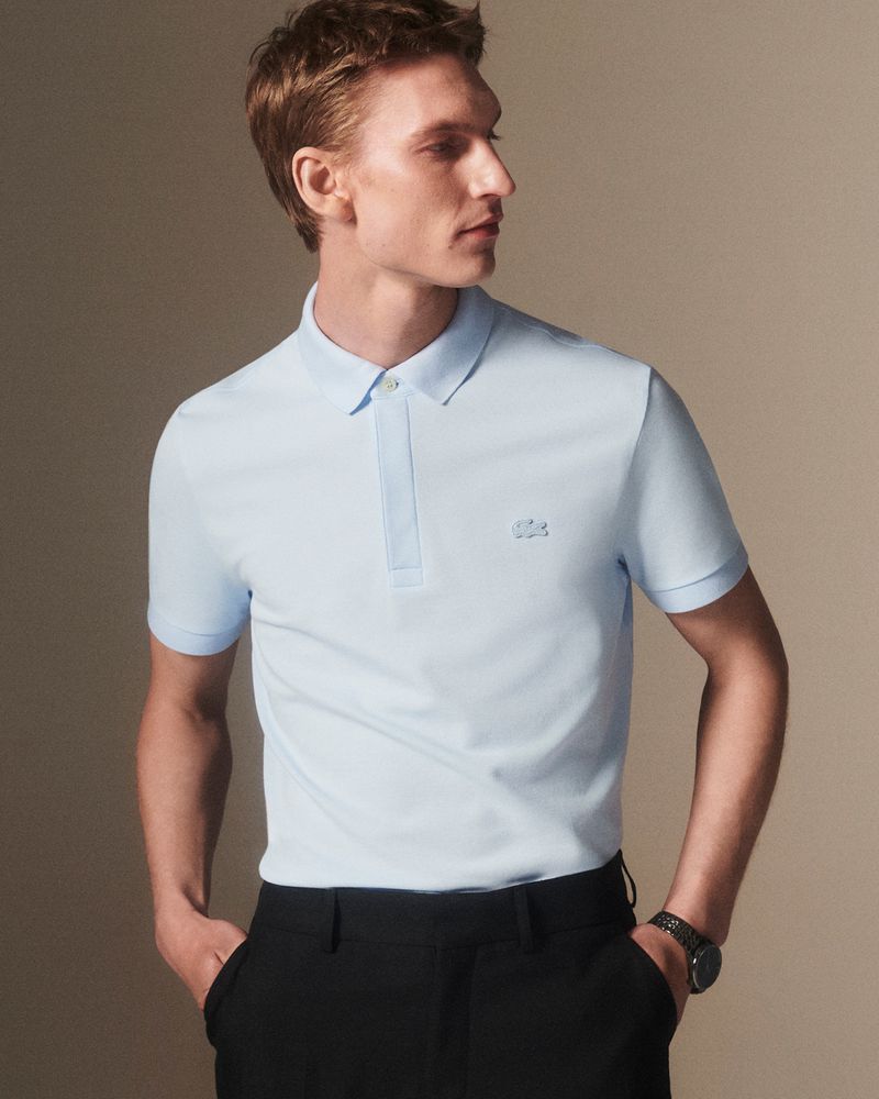 A fair-skinned adult male model poses indoors wearing a light blue polo shirt and black dress pants, his hands tucked in his pockets.