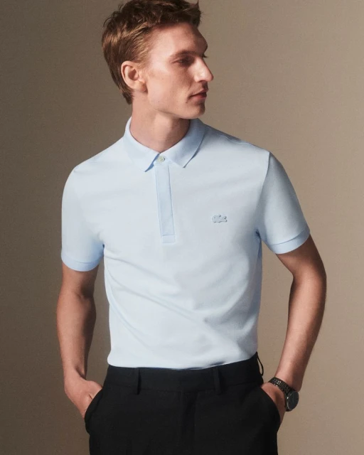 A fair-skinned adult male model poses indoors wearing a light blue polo shirt and black dress pants, his hands tucked in his pockets.