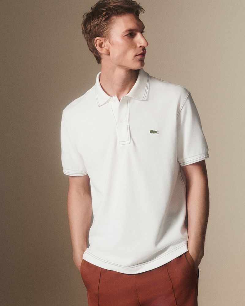 Blond male model wears a white Lacoste polo shirt and rust-colored pants against a neutral studio backdrop for a fashion advertisement.