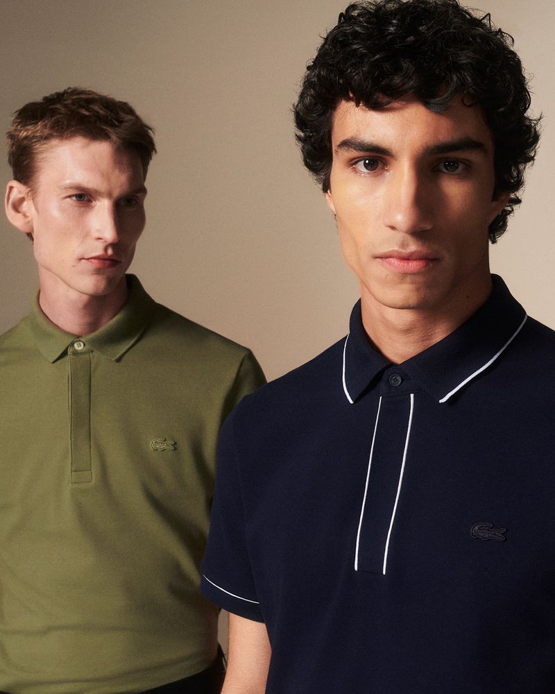 Two adult men pose indoors, one in a green polo, the other in a navy polo shirt with white trim, against a neutral background in a studio setting.