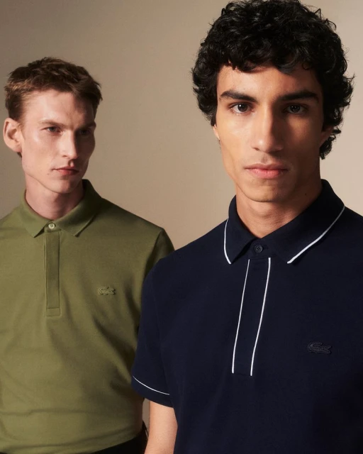 Two adult men pose indoors, one in a green polo, the other in a navy polo shirt with white trim, against a neutral background in a studio setting.