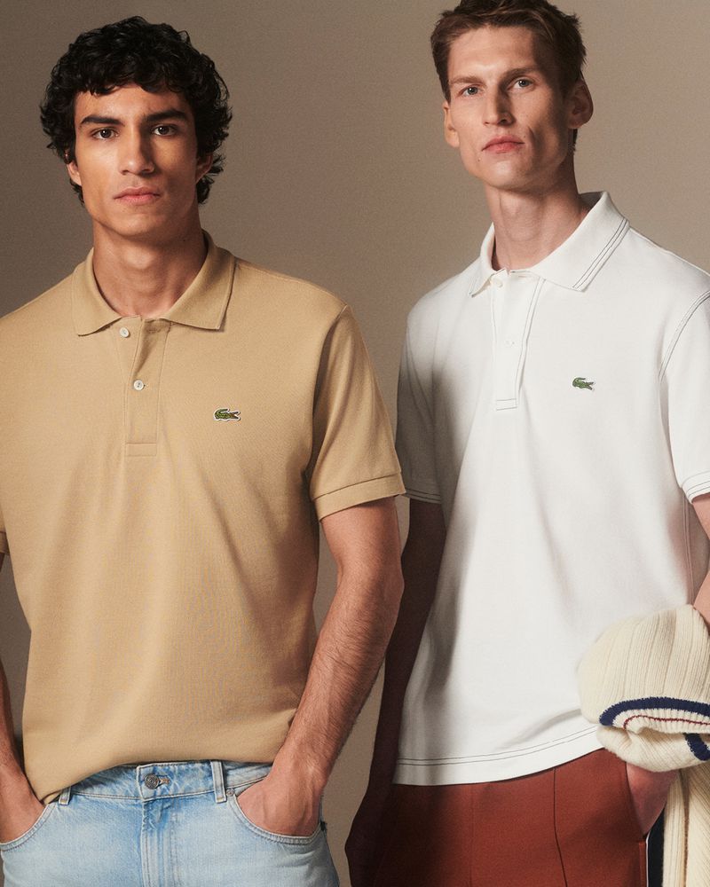 Two adult males wearing polo shirts by Lacoste, one in tan with light blue jeans and the other in white with brown pants.