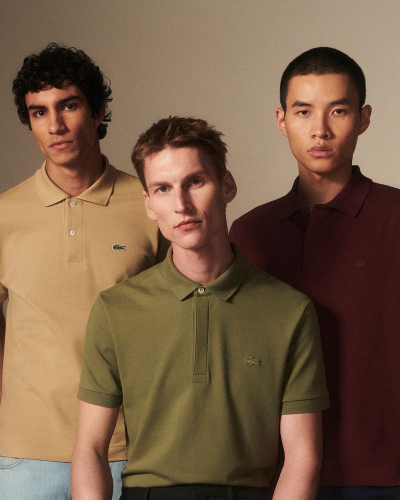 Three adult male models in solid color Lacoste polo shirts pose indoors against a neutral background, showcasing a fashion editorial.