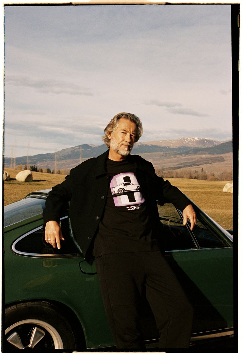 Mature man in black jacket and tee-shirt leans on a green car in front of a rural landscape with rolling hills and sky.