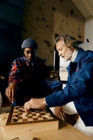Two men play checkers indoors at a wooden table. The man closest to the camera reaches out to move a game piece.