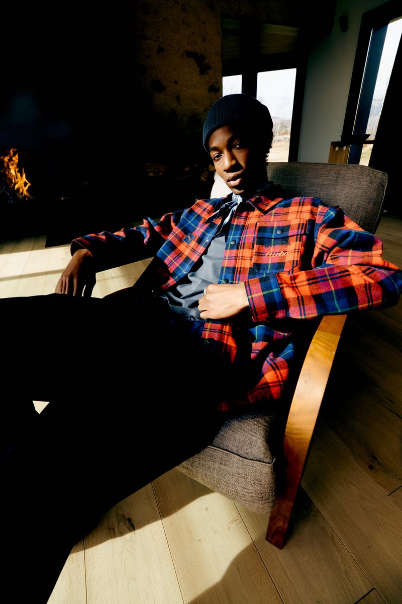 A young adult male, wearing a beanie and plaid shirt, is sitting in an armchair in a sunlit room with a fireplace.