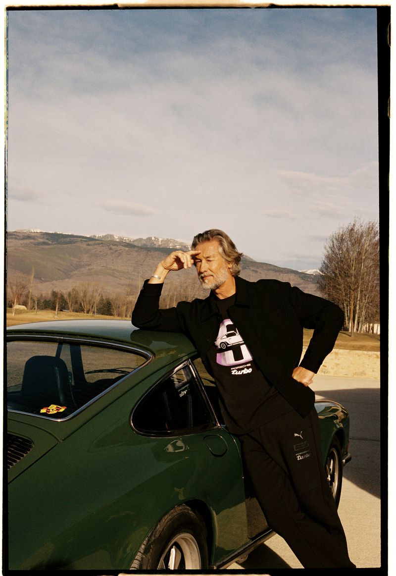 Mature man leans against a vintage green Porsche in an outdoor setting with mountains and sky visible in the background.