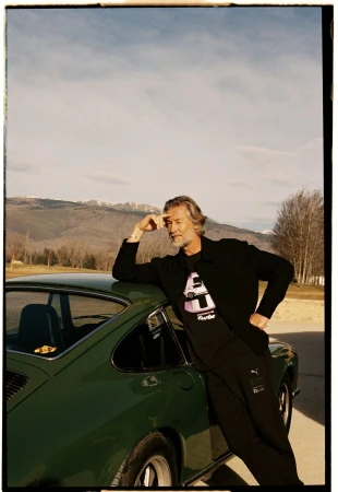 Mature man leans against a vintage green Porsche in an outdoor setting with mountains and sky visible in the background.