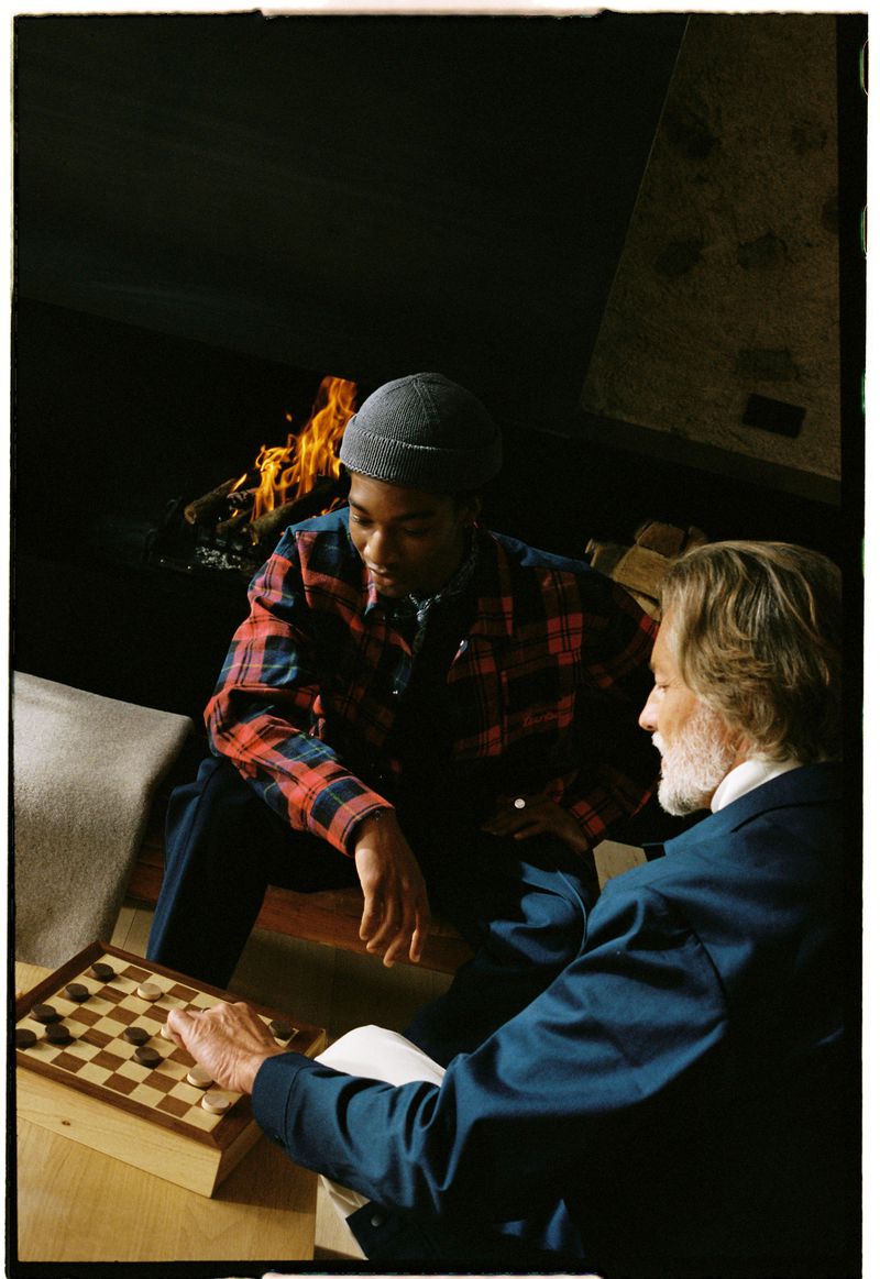 Two men play checkers indoors near a fireplace. One man wears a blue jacket and the other wears a red and blue plaid shirt.