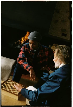 Two men play checkers indoors near a fireplace. One man wears a blue jacket and the other wears a red and blue plaid shirt.