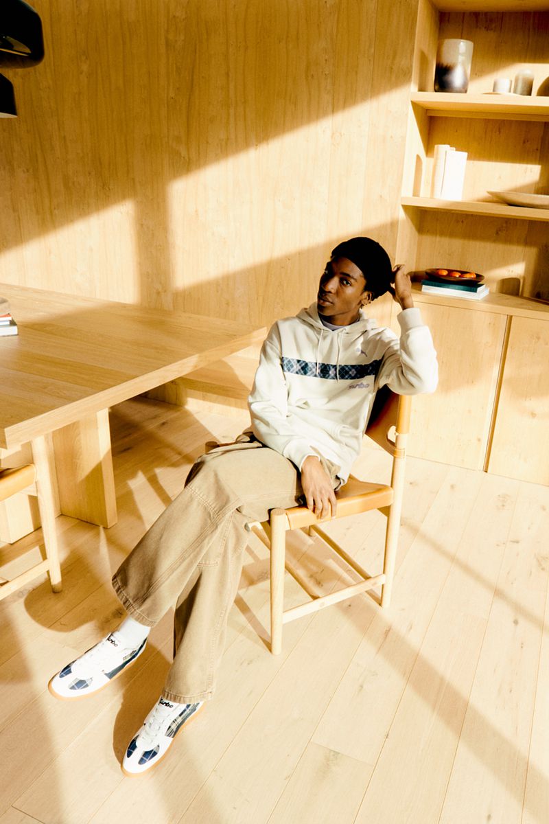 A young Black man seated on a wooden chair in a sunlit room, wearing a patterned white hoodie, beige trousers, and sneakers.