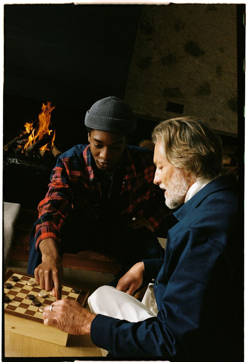 Two men of different ages play checkers next to a fireplace. The younger man is wearing a gray hat and red flannel shirt.