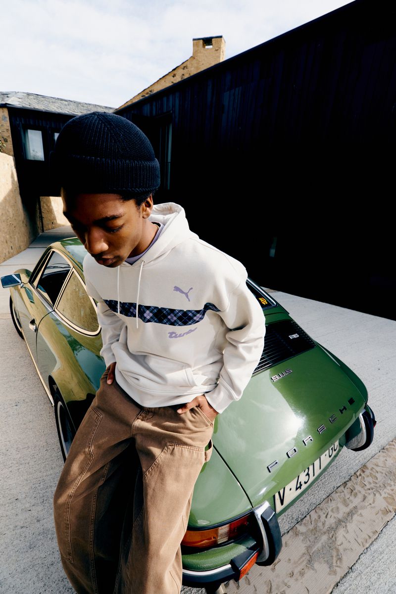 A young Black man in a hoodie leans against a vintage green Porsche. He's wearing a black beanie and tan pants in an outdoor shot.