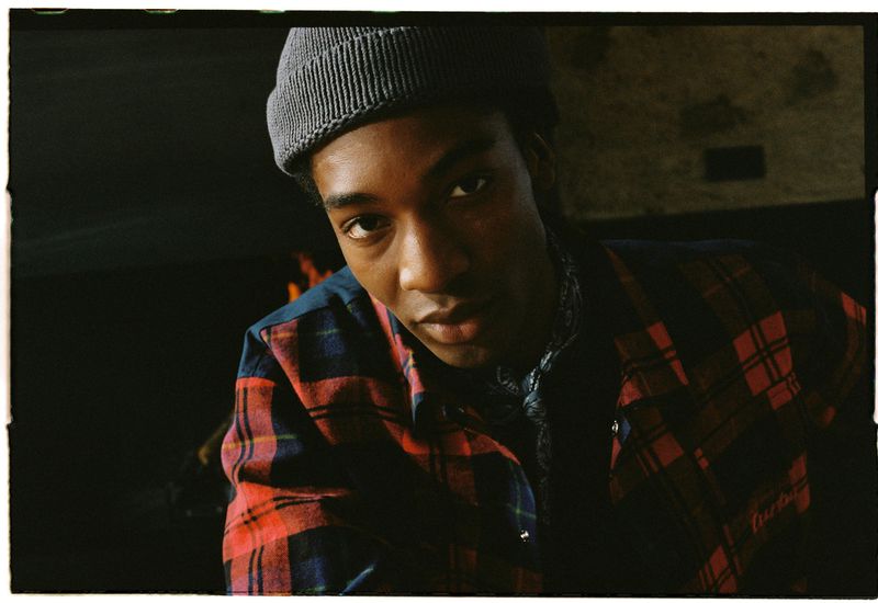Close-up of a Black male model wearing a gray beanie and a red and blue plaid shirt, with a dark background and soft lighting.