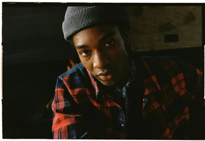 Close-up of a Black male model wearing a gray beanie and a red and blue plaid shirt, with a dark background and soft lighting.