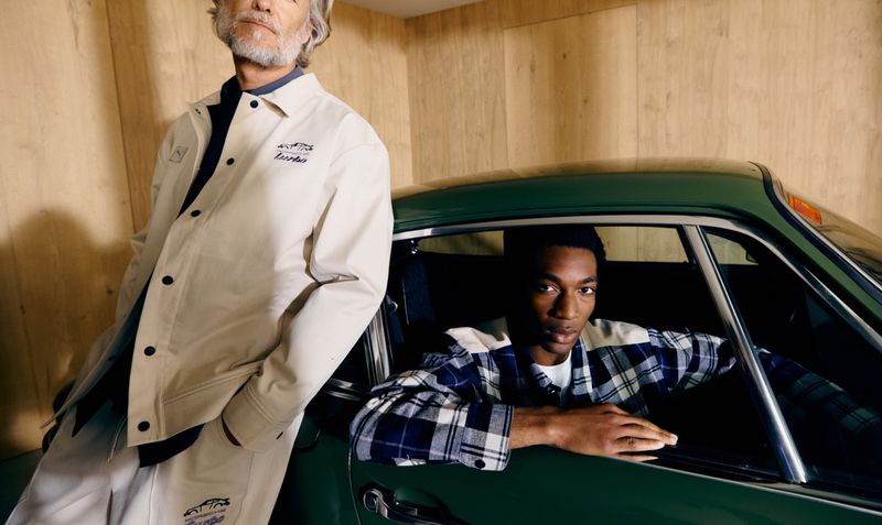 Two men pose indoors with a green classic car. One man sits inside the car, while the other stands beside it against a wood wall.