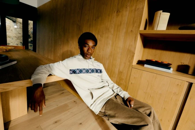 Young Black man wearing a white hoodie and beanie seated indoors near a wooden table against wood paneling