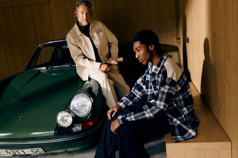 Two men pose indoors with a vintage green Porsche. One man sits on a bench, the other leans on the car in casual attire.
