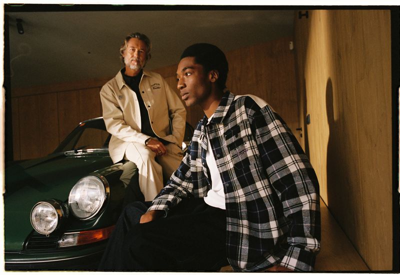 Two men pose with a green sports car inside of a garage. One man is seated on the car's hood, wearing a tan jacket. The other stands.
