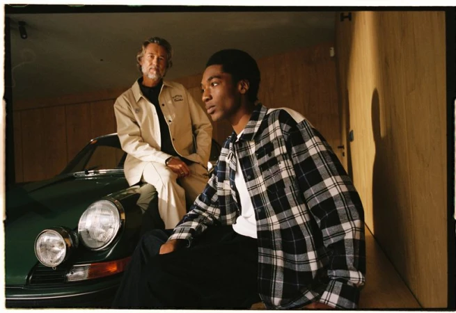 Two men pose with a green sports car inside of a garage. One man is seated on the car's hood, wearing a tan jacket. The other stands.