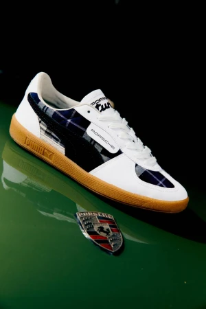 Puma x Porsche trainers on a green surface with the Porsche emblem in an eye-level, medium studio shot with dark background.