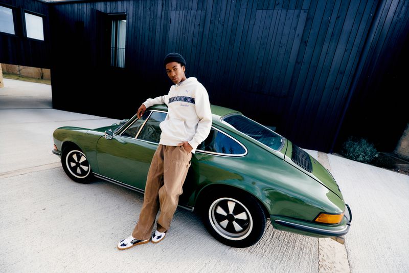 A young Black man leans casually against a classic green Porsche parked in front of a modern, dark-wood building on a sunny day.