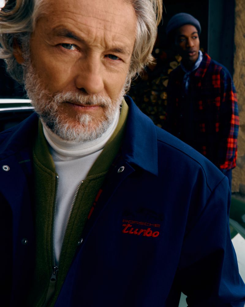 Older Caucasian man with a beard and blue jacket in medium shot, with a younger Black man in the background and a floral design.