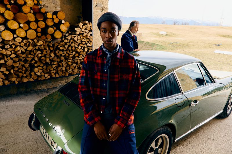 Portrait of a stylish young adult in a plaid shirt leaning against a vintage Porsche 911, with an older adult in the background.