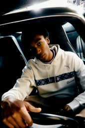 Young Black man in a white Puma hoodie seated behind the steering wheel of a Porsche sports car